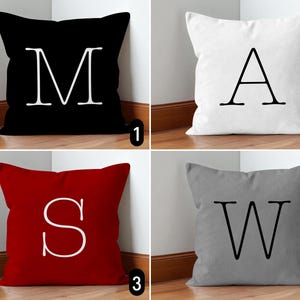 May include: Four square throw pillows in black, white, red, and gray. Each pillow features a large, white letter: M, A, S, and W. The pillows are leaning against a white wall and a wooden floor.
