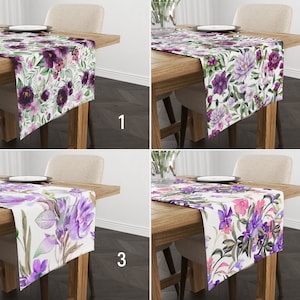 May include: Four floral table runners with different designs are displayed on wooden tables. Each runner features a white background with a variety of purple, pink, and green floral patterns. The runners are rectangular and appear to be made of fabric.