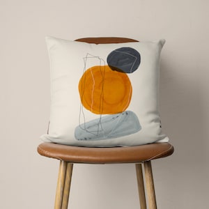 Puede incluir: Una almohada blanca con un dise&ntilde;o abstracto que presenta un c&iacute;rculo amarillo, un c&iacute;rculo azul y una forma gris. El dise&ntilde;o recuerda a una pintura de acuarela.
