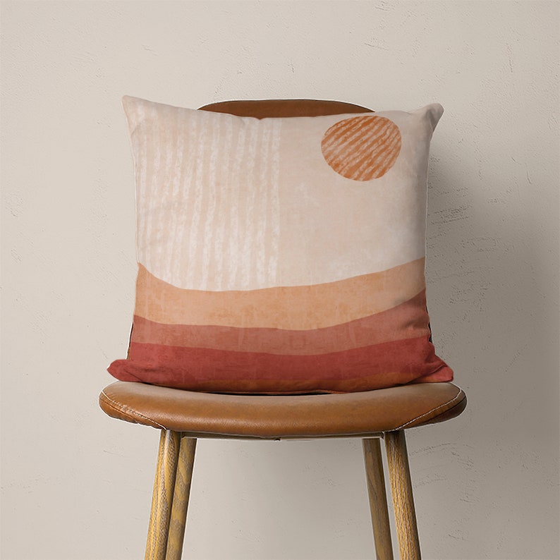May include: A throw pillow with a geometric design featuring a sun, stripes, and a layered landscape in shades of orange, brown, and red. The pillow is sitting on a brown leather chair.