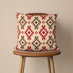 Op de afbeelding: Een decoratief kussen met een geometrisch patroon in tinten rood, bruin en cr&egrave;me. Het kussen ligt op een bruine houten stoel.