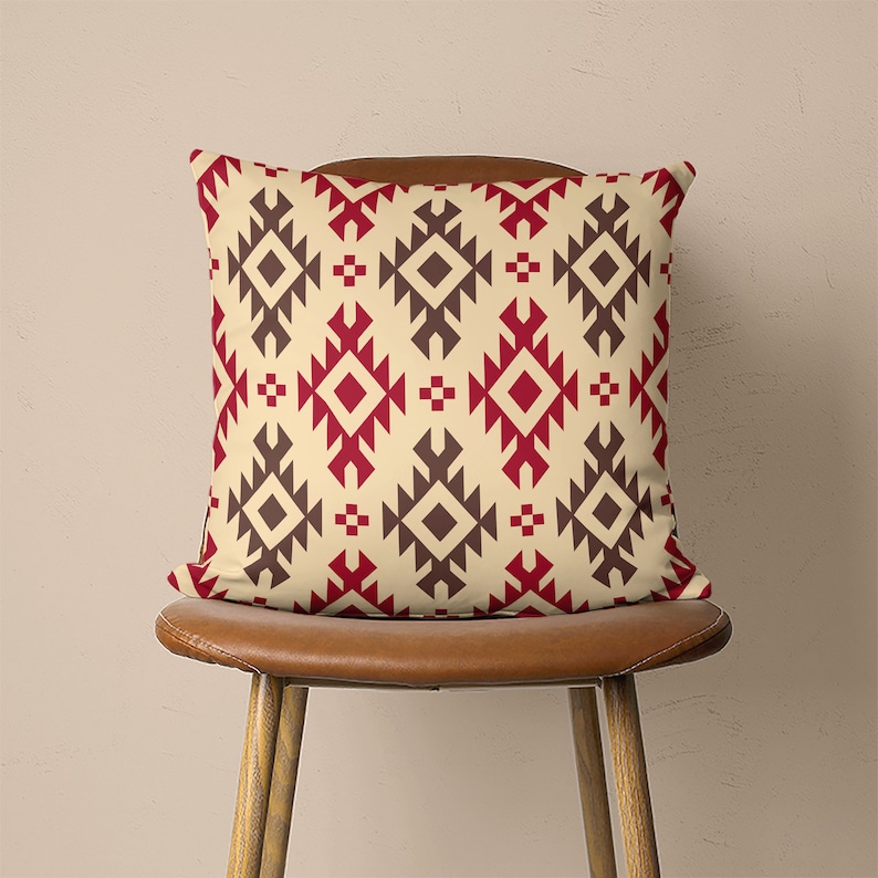 Op de afbeelding: Een decoratief kussen met een geometrisch patroon in tinten rood, bruin en cr&egrave;me. Het kussen ligt op een bruine houten stoel.