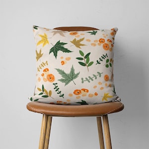 Puede incluir: Una almohada de tiro con un fondo de color crema y un patr&oacute;n de hojas verdes, flores naranjas y hojas de arce amarillas.