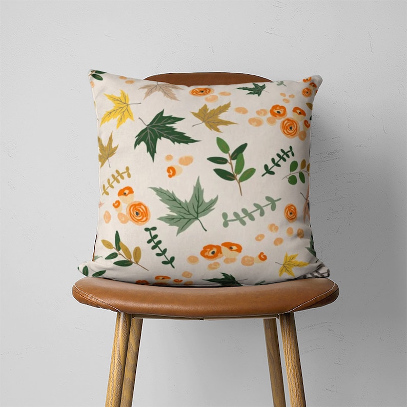 Puede incluir: Una almohada de tiro con un fondo de color crema y un patr&oacute;n de hojas verdes, flores naranjas y hojas de arce amarillas.