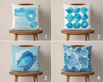 Könnte beinhalten: Vier verschiedene Kissen-Designs mit blauen Aquarellmustern. Die Kissen befinden sich auf braunen Stühlen mit hellen Holzbeinen. Die Kissen sind von 1 bis 4 nummeriert.