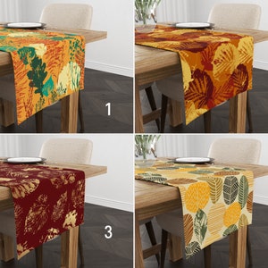 May include: Four different table runners with autumn leaf designs. The runners are in various colors, including orange, yellow, green, and brown. The runners are all on wooden tables with chairs.