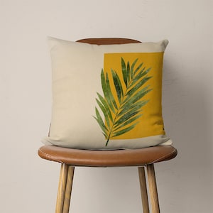 May include: A throw pillow with a cream-colored background and a yellow square with a green palm leaf design.
