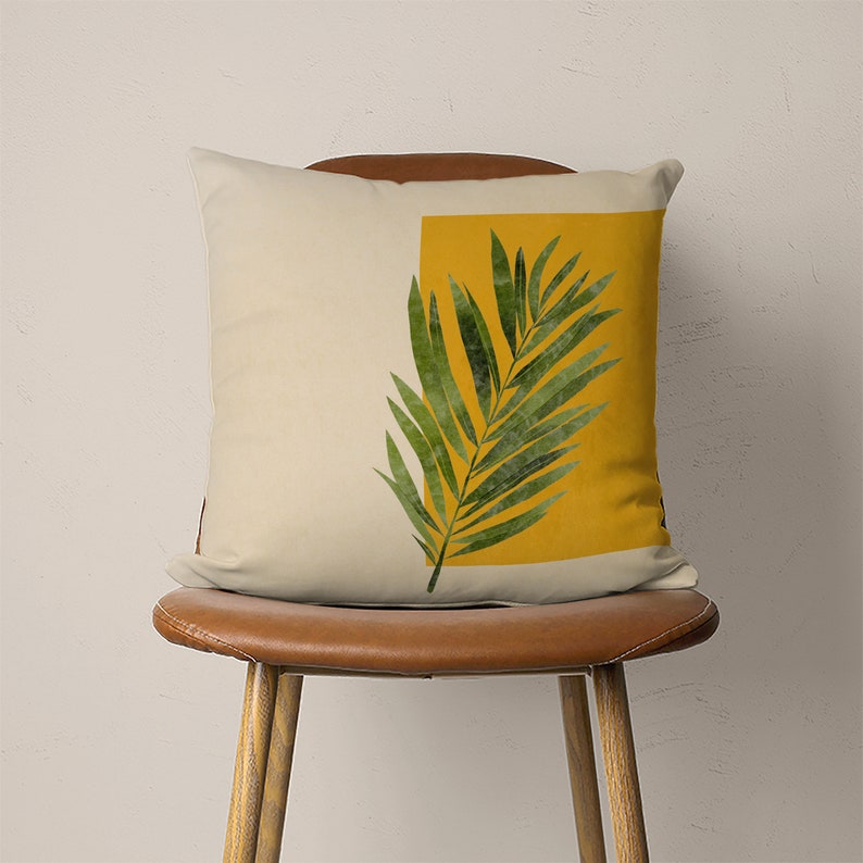 May include: A throw pillow with a cream-colored background and a yellow square with a green palm leaf design.