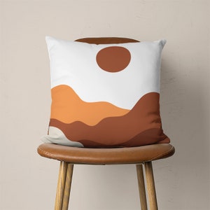 Pu&ograve; includere: Un cuscino bianco con un sole marrone e un design astratto di montagna arancione e marrone.