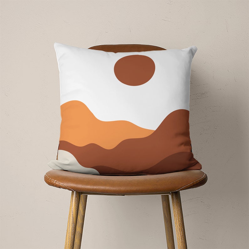 Pu&ograve; includere: Un cuscino bianco con un sole marrone e un design astratto di montagna arancione e marrone.