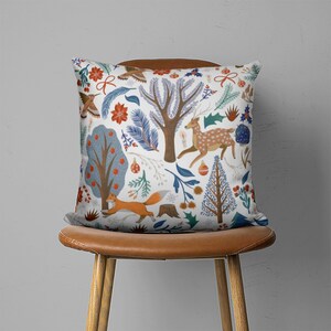 Puede incluir: Una almohada blanca con un patr&oacute;n colorido de &aacute;rboles, ciervos, zorros y p&aacute;jaros. El patr&oacute;n recuerda a una escena de bosque de invierno.