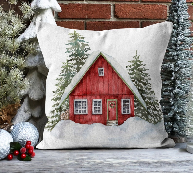 May include: A white throw pillow with a watercolor illustration of a red cabin in a snowy forest. The cabin has a wreath on the door and snow on the roof.