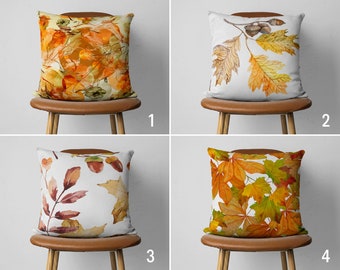 Può includere: Quattro cuscini decorativi con diversi motivi di foglie autunnali. I cuscini sono posizionati su sedie marroni su uno sfondo bianco. I cuscini sono tutti quadrati e in tessuto bianco con motivi di foglie di diversi colori.
