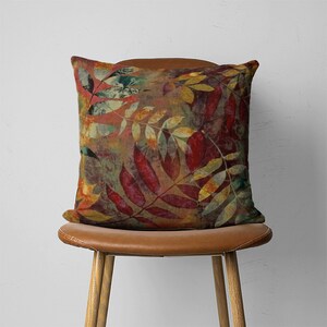Puede incluir: Una almohada cuadrada con un estampado abstracto de hojas de colores en tonos rojos, verdes, amarillos y marrones. La almohada est&aacute; sentada en una silla de cuero marr&oacute;n.