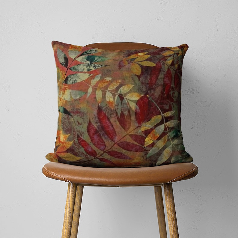 Puede incluir: Una almohada cuadrada con un estampado abstracto de hojas de colores en tonos rojos, verdes, amarillos y marrones. La almohada est&aacute; sentada en una silla de cuero marr&oacute;n.