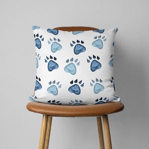 May include: A white square pillow with a repeating pattern of blue paw prints. The paw prints vary in shades of blue, creating a watercolor effect. The pillow is placed on a brown leather chair.