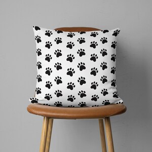 May include: A square white pillow with a repeating pattern of black paw prints. The pillow is placed on a brown leather chair with wooden legs. The background is a plain gray wall.