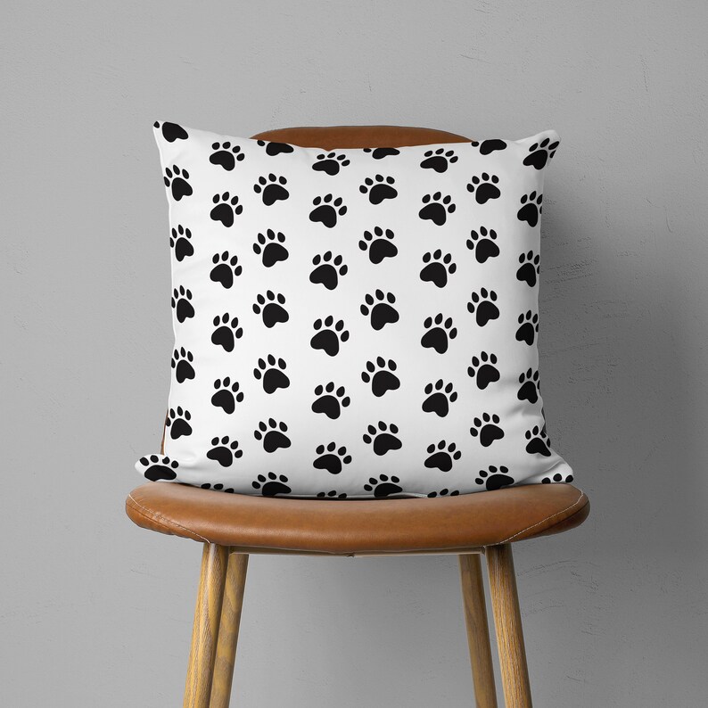 May include: A square white pillow with a repeating pattern of black paw prints. The pillow is placed on a brown leather chair with wooden legs. The background is a plain gray wall.