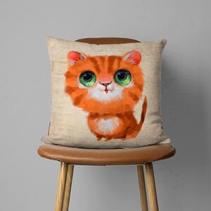 May include: A throw pillow with a cartoon illustration of an orange tabby cat with big green eyes. The cat is sitting on a beige background.