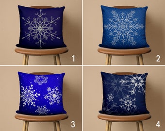 May include: Four blue throw pillows with white snowflake designs. The pillows are on brown chairs with a light brown background.