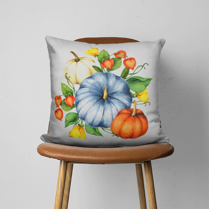 May include: A white throw pillow with a blue, white, and orange pumpkin design. The pumpkins are surrounded by green leaves and orange flowers.