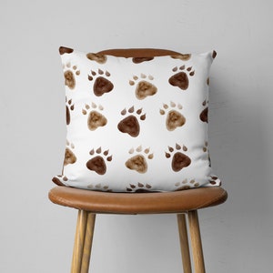 May include: A square decorative pillow with a white background and a repeating pattern of brown paw prints. The pillow is displayed on a brown leather chair, showcasing its design and texture.