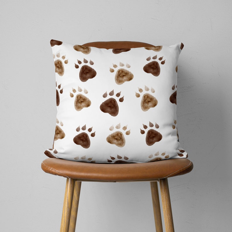 May include: A square decorative pillow with a white background and a repeating pattern of brown paw prints. The pillow is displayed on a brown leather chair, showcasing its design and texture.