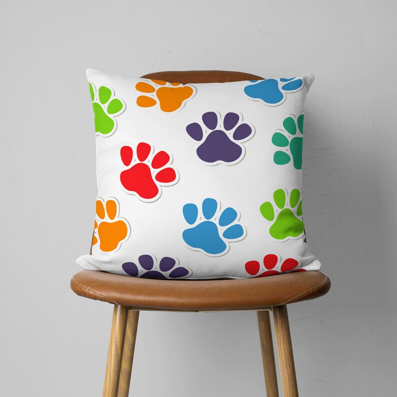 May include: A white square pillow with a repeating pattern of colorful paw prints in red, orange, purple, blue, and green. The pillow is on a brown leather chair with wooden legs. The paw prints have a white outline.