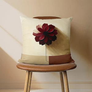 May include: A beige pillow with a red and white flower design. The pillow is sitting on a brown leather chair.