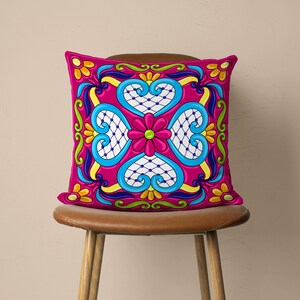 May include: A pink cushion with a colourful Mexican-style tile pattern featuring blue, yellow, green, and white hearts and flowers.
