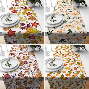 Pode incluir: Quatro caminhos de mesa diferentes com designs de folhas de outono. Os caminhos de mesa são brancos com folhas de várias cores, incluindo vermelho, laranja, amarelo e verde. Alguns caminhos de mesa apresentam bolotas e bagas.