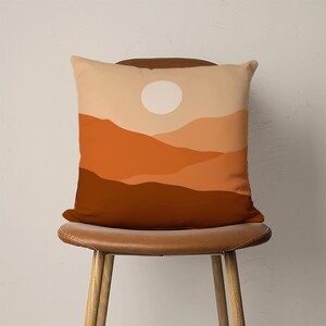 Pu&ograve; includere: Un cuscino decorativo con un design minimalista che presenta un cerchio bianco su un paesaggio di montagna stratificato in arancione e marrone.