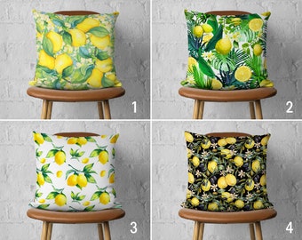 May include: Four throw pillows with different lemon designs. The pillows are on brown chairs against a white brick wall. The first pillow has a yellow lemon design on a white background. The second pillow has a green tropical leaf design with yellow lemons. The third pillow has a white background with a yellow lemon design. The fourth pillow has a black background with a yellow lemon design.
