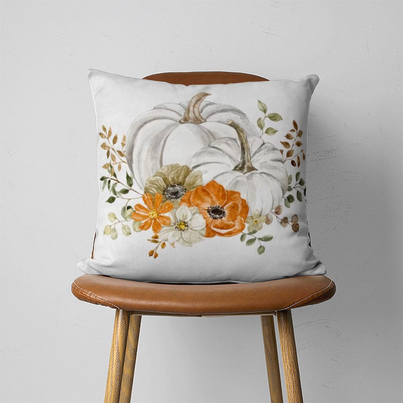 May include: White pumpkins with orange and white flowers on a white pillow. The pillow is on a brown chair.