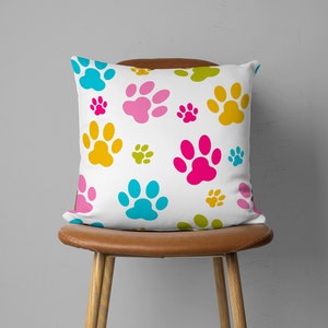May include: A square white pillow with a repeating pattern of colorful paw prints in shades of blue, pink, yellow, and green. The pillow is placed on a brown leather chair, creating a playful and vibrant aesthetic.