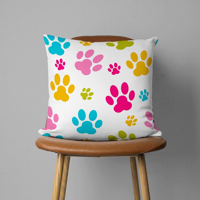 May include: A square white pillow with a repeating pattern of colorful paw prints in shades of blue, pink, yellow, and green. The pillow is placed on a brown leather chair, creating a playful and vibrant aesthetic.