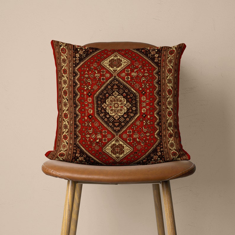 May include: A red, black and cream throw pillow with a traditional Persian rug pattern. The pillow is sitting on a brown leather chair.