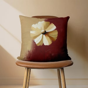 May include: A decorative throw pillow with a beige and burgundy background featuring a large, white and gold flower with a brown stem.