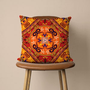 Op de afbeelding: Een oranje, rood en blauw gepatroneerd kussen met een mandala-ontwerp. Het kussen ligt op een bruine stoel.