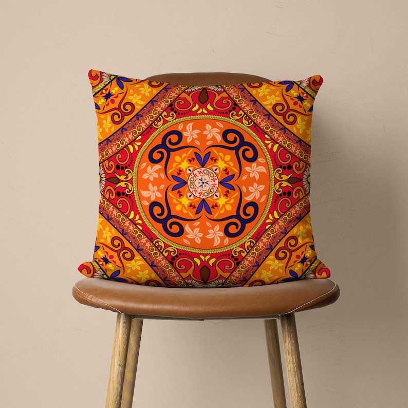 Op de afbeelding: Een oranje, rood en blauw gepatroneerd kussen met een mandala-ontwerp. Het kussen ligt op een bruine stoel.