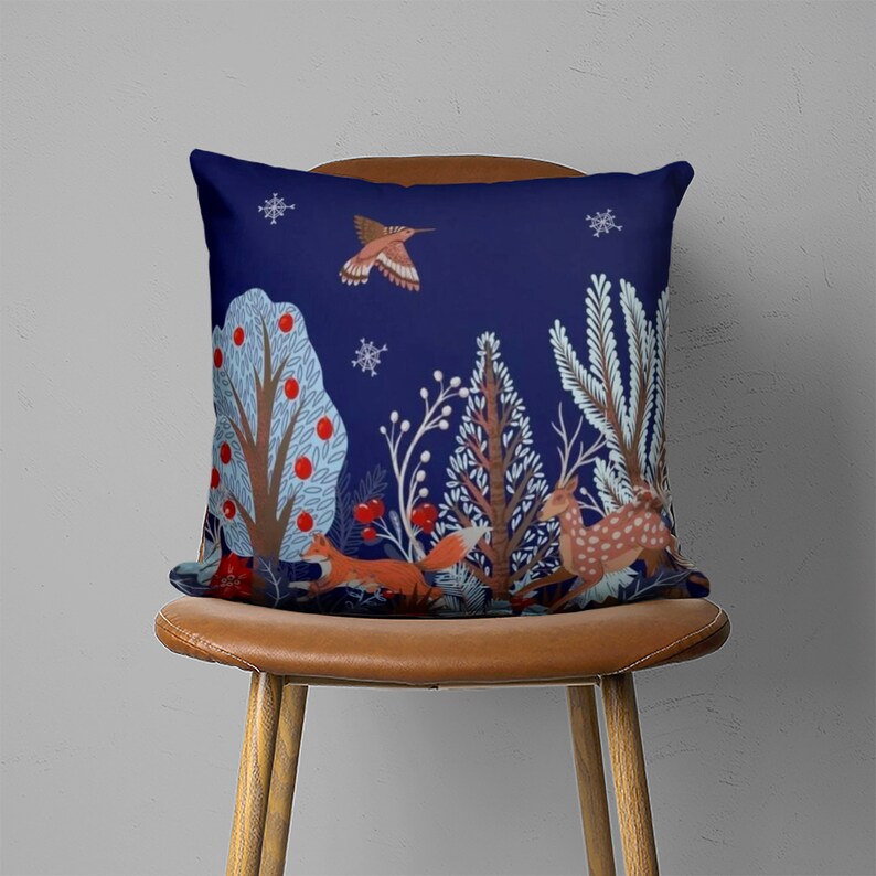 Puede incluir: Una almohada azul con una escena de bosque de invierno con un colibr&iacute;, un ciervo, zorros y &aacute;rboles con bayas rojas. La almohada est&aacute; en una silla marr&oacute;n.