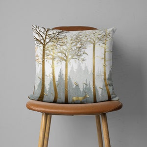 May include: A throw pillow with a white background and a gold metallic design of trees and a deer. The pillow is sitting on a brown leather chair.