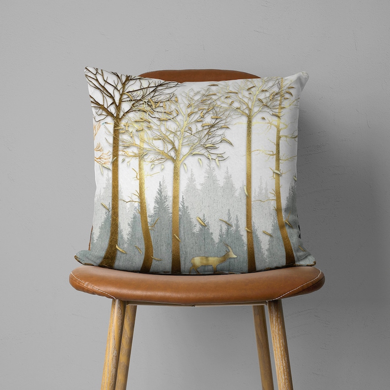 May include: A throw pillow with a white background and a gold metallic design of trees and a deer. The pillow is sitting on a brown leather chair.