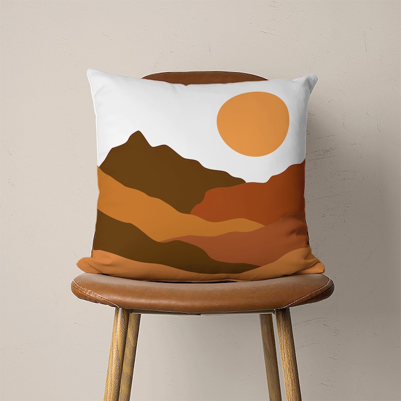 Pu&ograve; includere: Un cuscino bianco con un design astratto di paesaggio montano marrone e arancione e un sole giallo.
