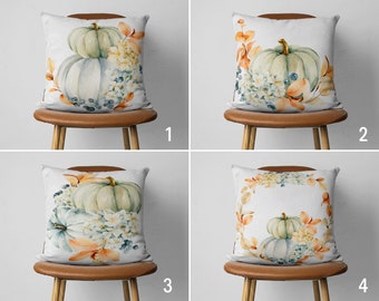 Könnte beinhalten: Vier Kissen mit Aquarellmotiven von Kürbissen, Blättern und Blumen. Die Kissen sind weiß mit hellblauem Hintergrund und auf braunen Stühlen angeordnet.