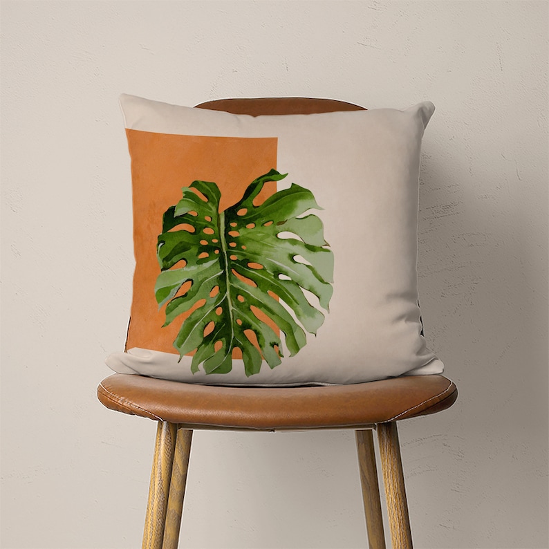 May include: A beige throw pillow with a green monstera leaf design on a burnt orange background.