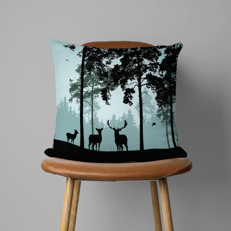 May include: A throw pillow with a blue and black design featuring a silhouette of a forest with deer. The deer are standing in a clearing in the forest. The pillow is on a brown chair.