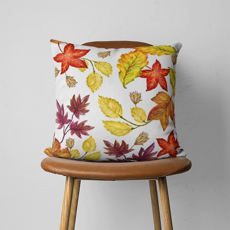 Puede incluir: Una almohada blanca con un estampado de hojas de oto&ntilde;o de colores. Las hojas son de color rojo, naranja, amarillo y marr&oacute;n. La almohada est&aacute; sentada en una silla de cuero marr&oacute;n.