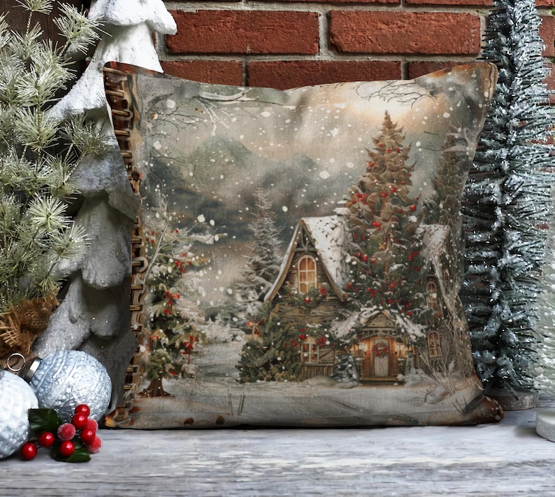 May include: A decorative throw pillow with a winter scene featuring a snow-covered cabin, a pine tree with red berries, and a snowy landscape. The pillow has a brown fabric with a distressed look and a zipper closure.