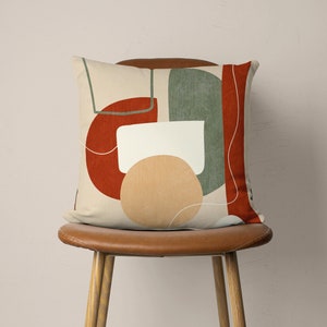 Puede incluir: Una almohada beige con un dise&ntilde;o geom&eacute;trico abstracto con formas rojas, verdes y beige. La almohada est&aacute; sentada en una silla de cuero marr&oacute;n.